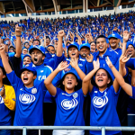 A vibrant scene capturing Salvadoran football fans cheering passionately in a stadium. People of various ages are present, all fully clothed in modest, casual attire, some wearing team colors. The atmosphere is electric with energy, showing diverse faces expressing joy and excitement. The stadium background is bustling but not overly detailed. This image should convey a strong sense of national pride and community spirit. Professional photography, high resolution, dynamic lighting, realistic, safe for work, appropriate content, fully clothed, family-friendly, perfect anatomy, correct proportions, natural pose, well-formed hands, proper finger count, natural body proportions.